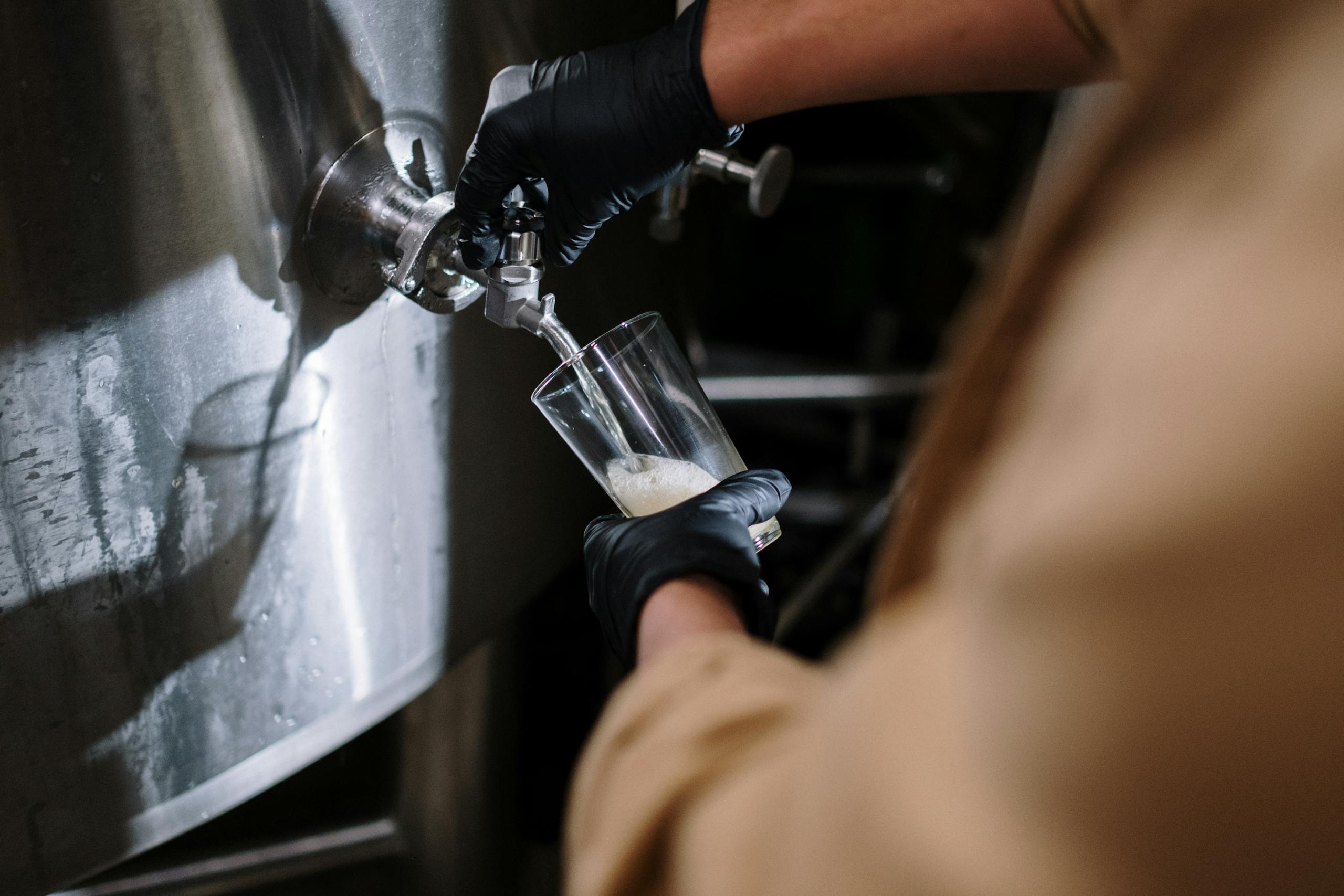 A gloved hand pours craft beer from a stainless steel tap at a brewery.