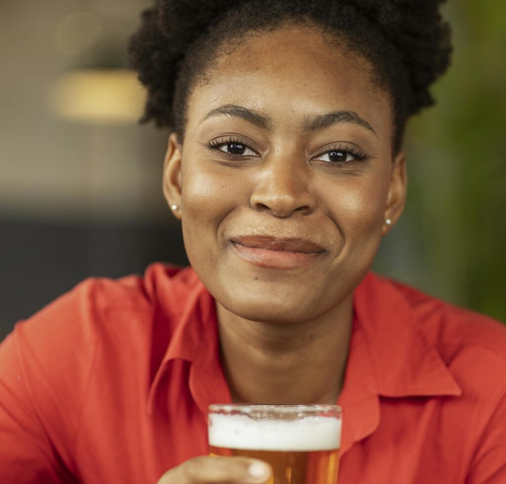 A cheerful woman enjoying a beer indoors, wearing a red shirt and smiling warmly.